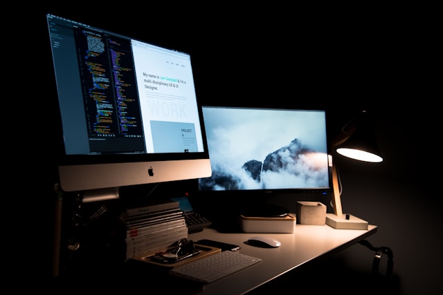 Designer's desk with iMac displaying code and UI, second monitor with landscape art
